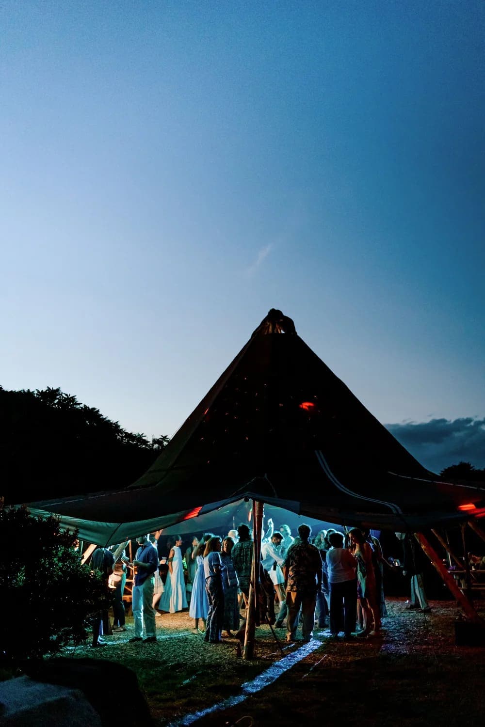 A large group of people dance and socialize under a tent at dusk.