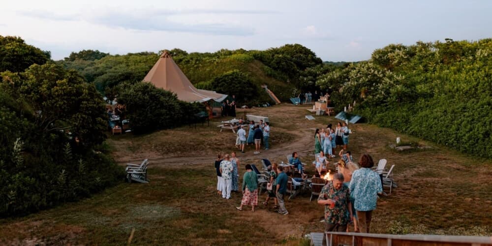 A lively outdoor gathering around a fire, with people socializing near a tent and lush greenery.
