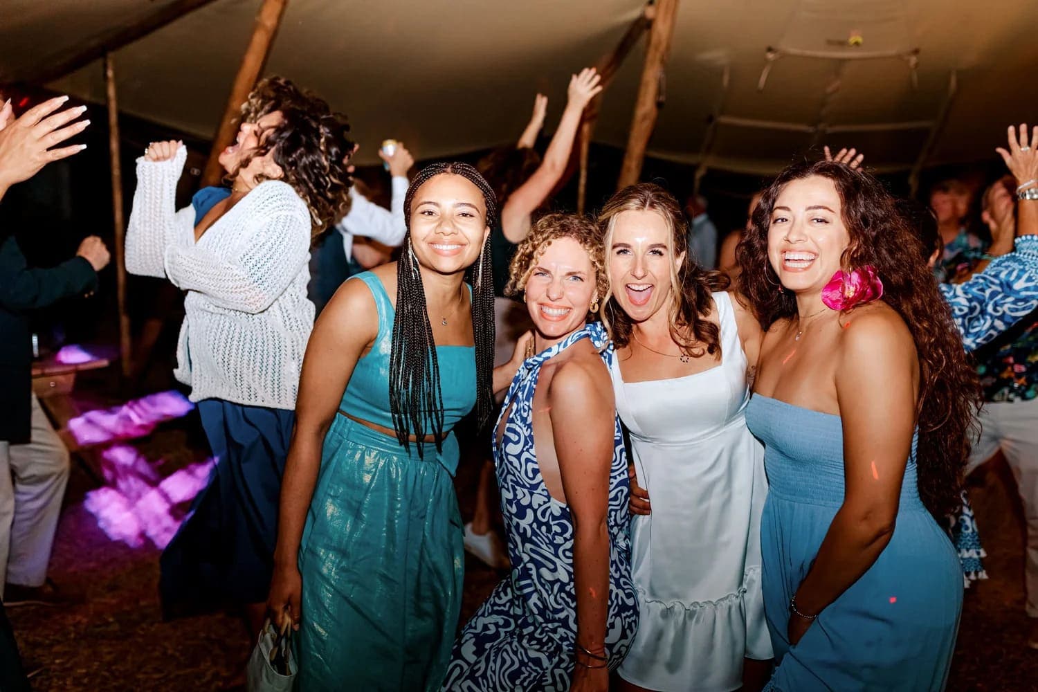 A group of four women smiling and posing together while others dance in the background.