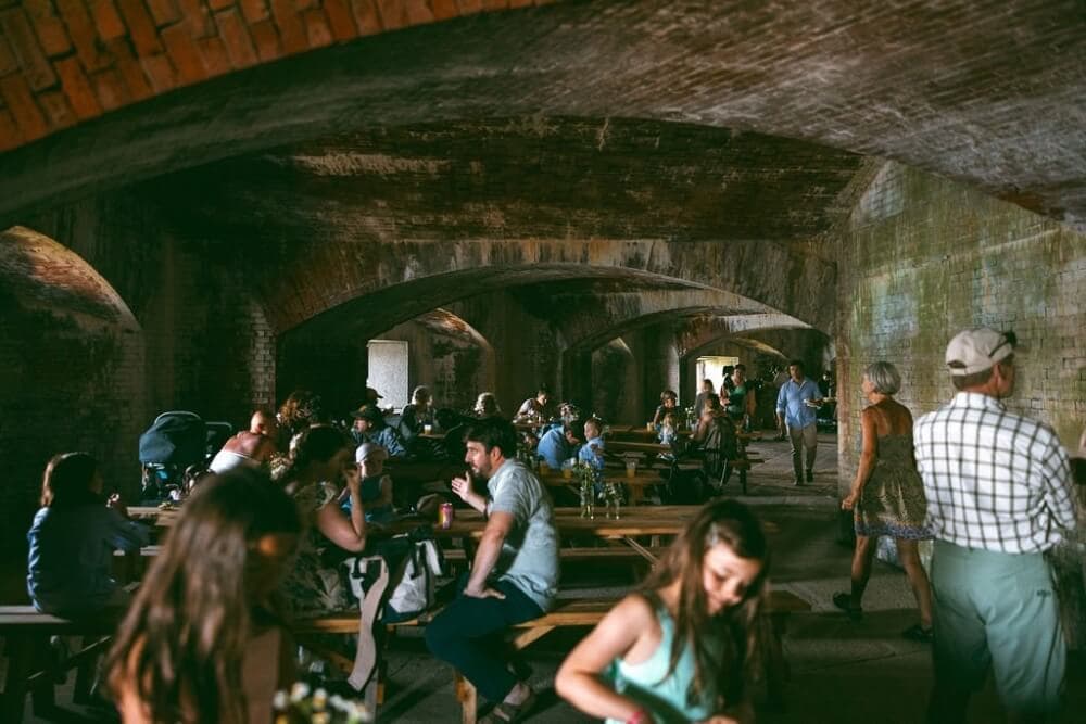 A crowded underground venue with people seated at long tables under arched brick ceilings.
