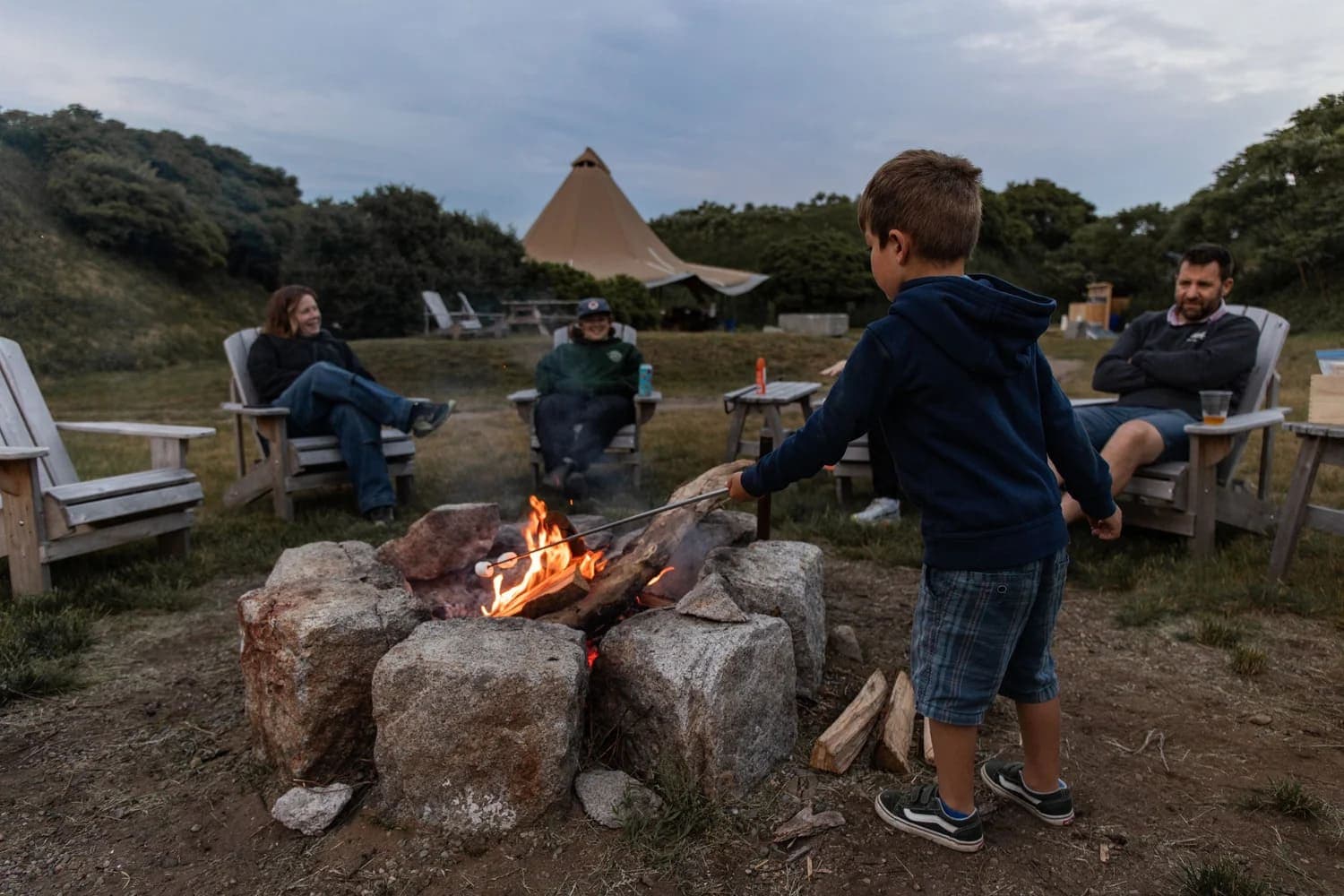 A young boy toasts a marshmallow over a campfire while adults relax nearby.