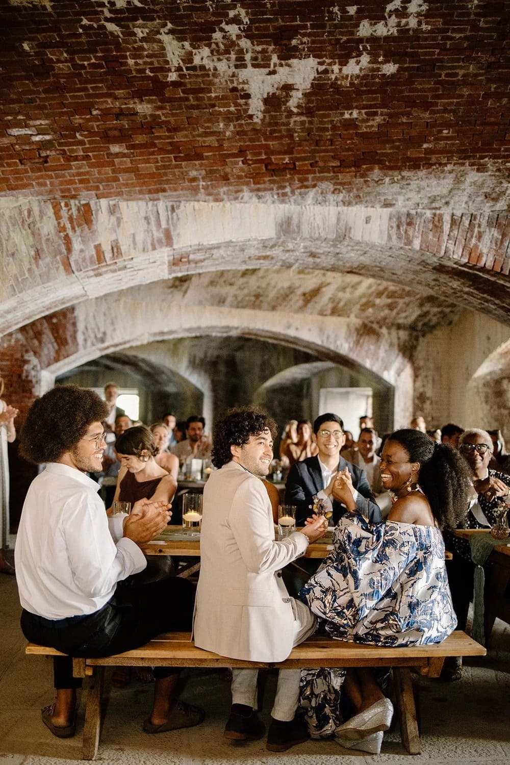 A diverse group of people seated at a long wooden table in a rustic, arched brick space, enjoying a lively gathering.