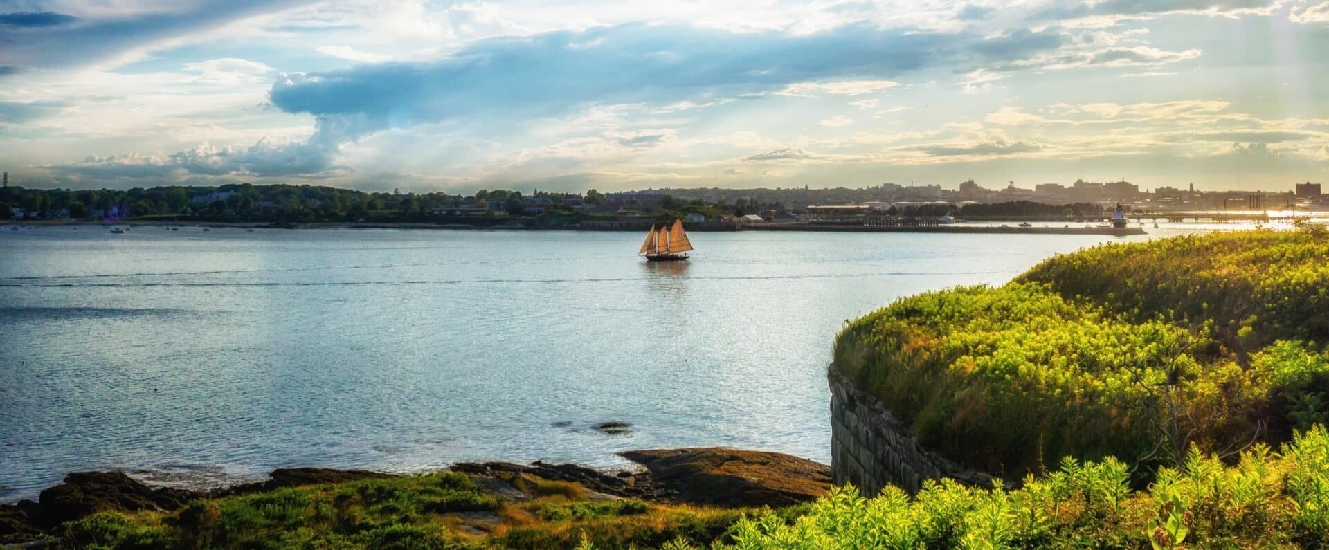 A sailing ship glides on calm waters near a lush, green shoreline under a partly cloudy sky.