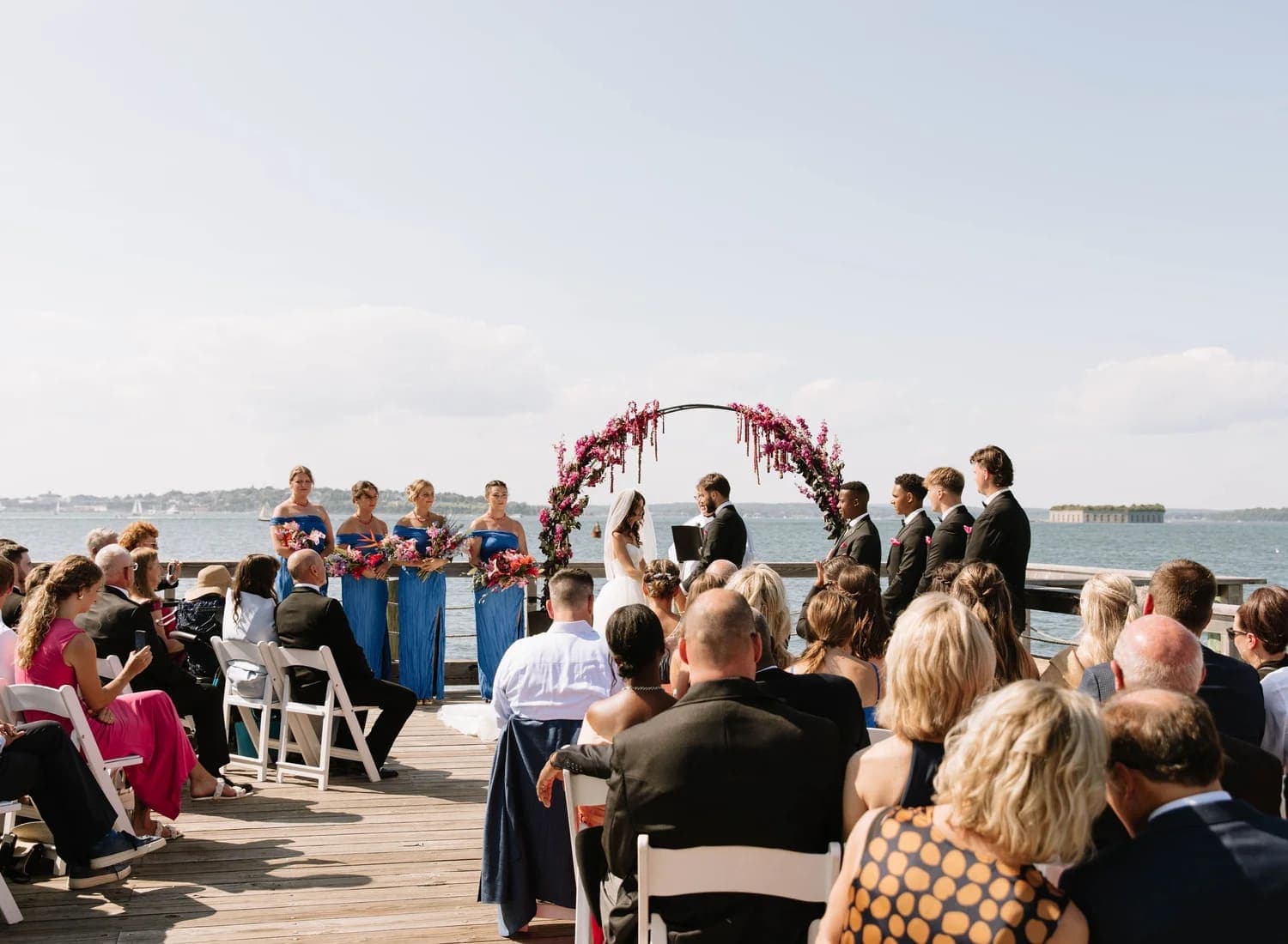 A wedding ceremony takes place by the waterfront with guests seated and the bridal party nearby.