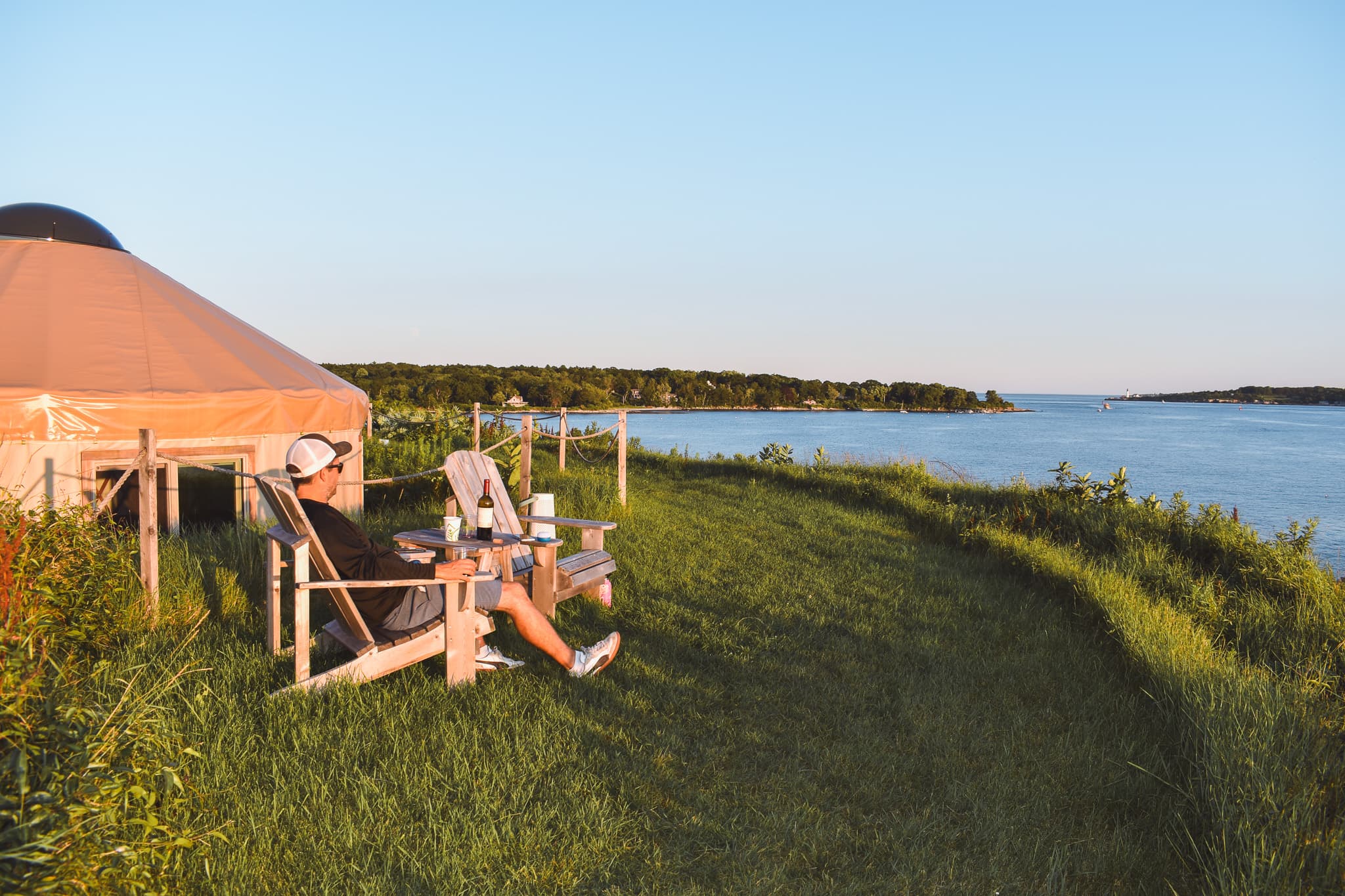 A person relaxes on a chair by a river, with a yurt in the background.