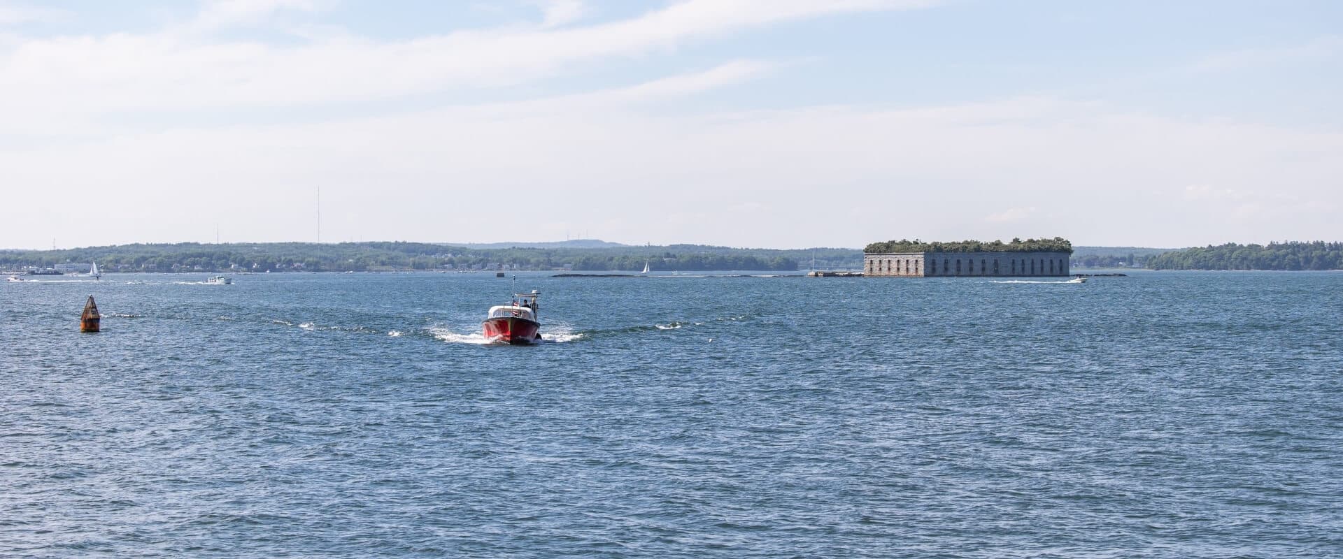 A small red boat navigates through blue waters near a distant structure on an island.