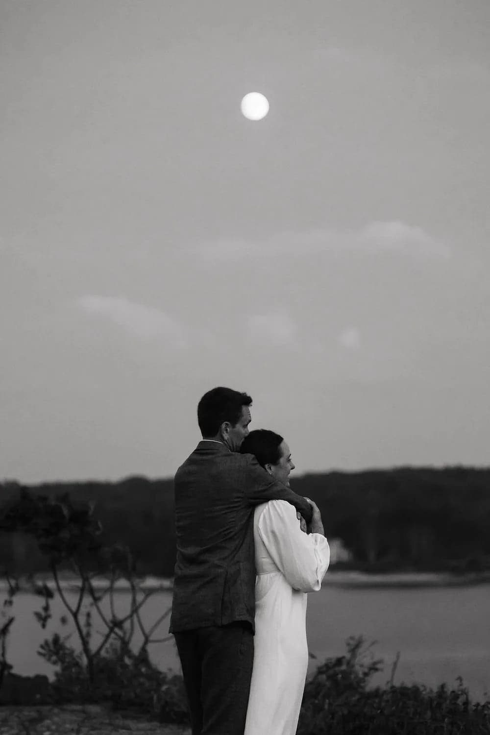 A couple embraces by a body of water under a full moon in a black and white setting.