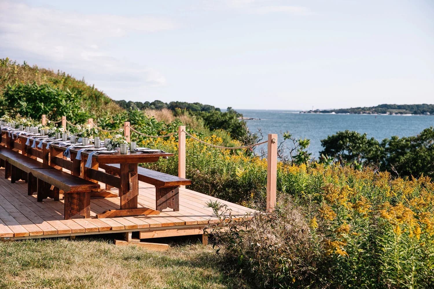 A wooden dining table set outdoors overlooking a river surrounded by greenery.