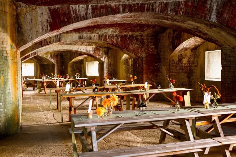 A rustic dining area with wooden tables adorned with floral centerpieces under arched brick ceilings.