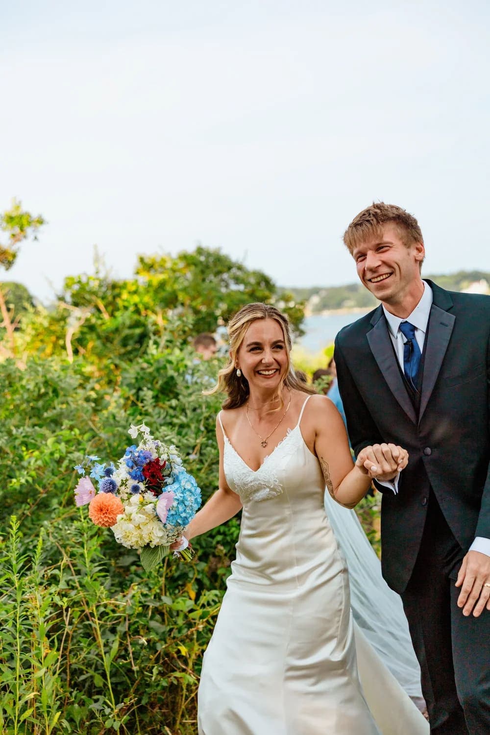 A happy couple walks hand in hand through a lush, green landscape, with the bride holding a colorful bouquet.