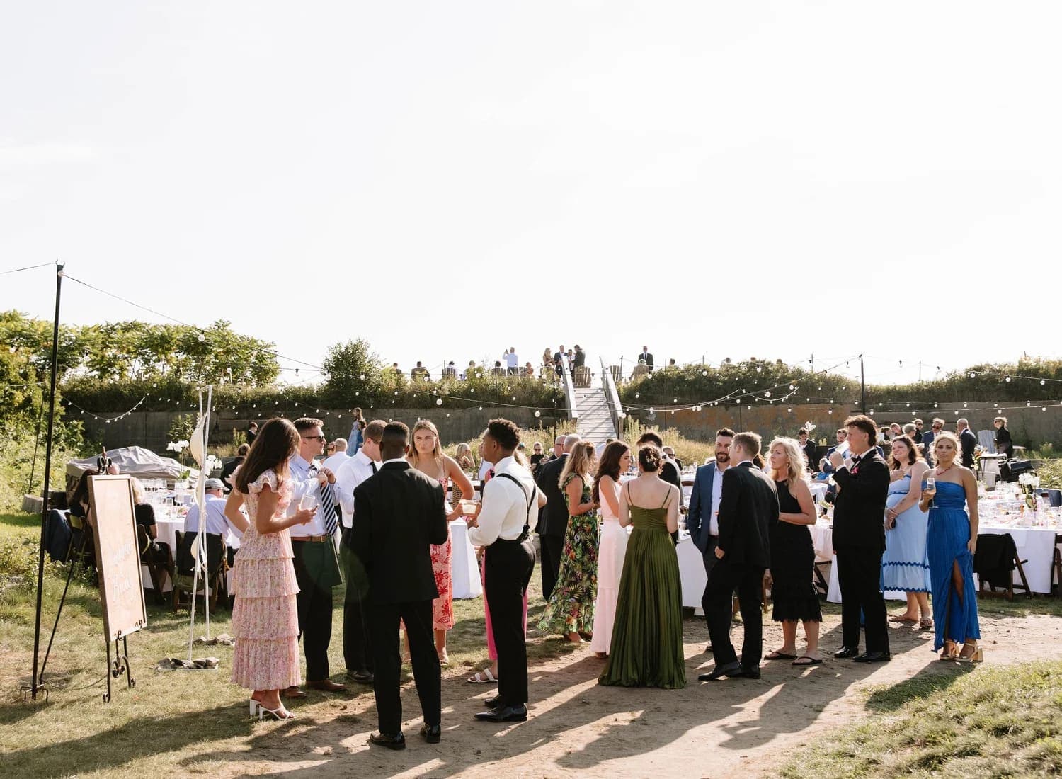 A group of elegantly dressed guests mingles at an outdoor event beneath a clear sky.