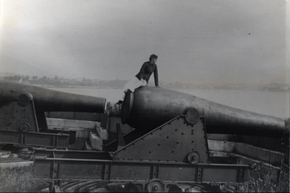 A man sits atop a large cannon near a body of water.
