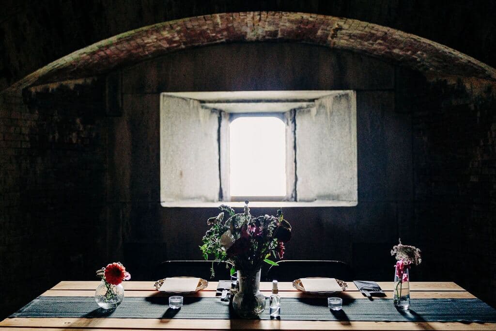 A wooden table set for a meal, adorned with flower vases, under a stone archway with a bright window.