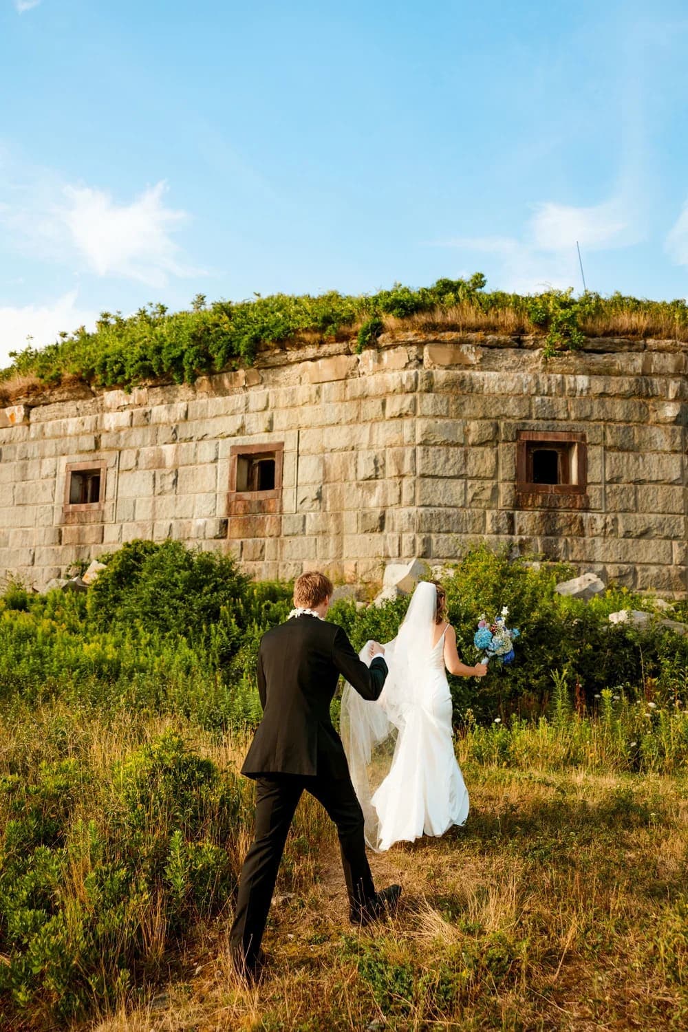 A couple in wedding attire walks hand in hand towards a historic stone structure.