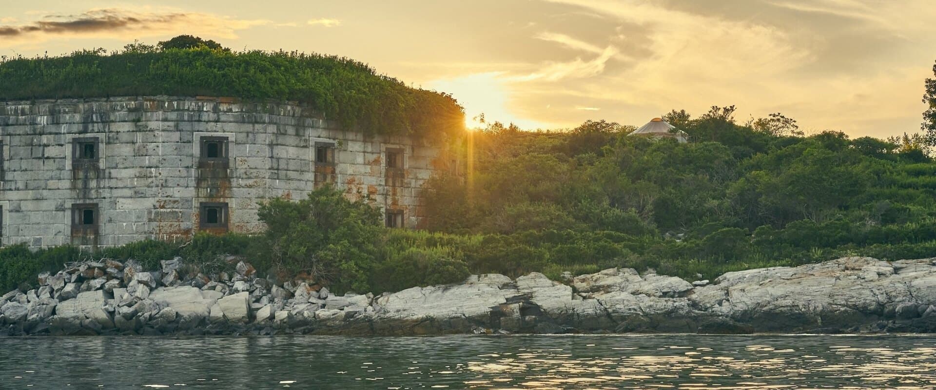 An old, overgrown stone building sits by the water with a sunset in the background.