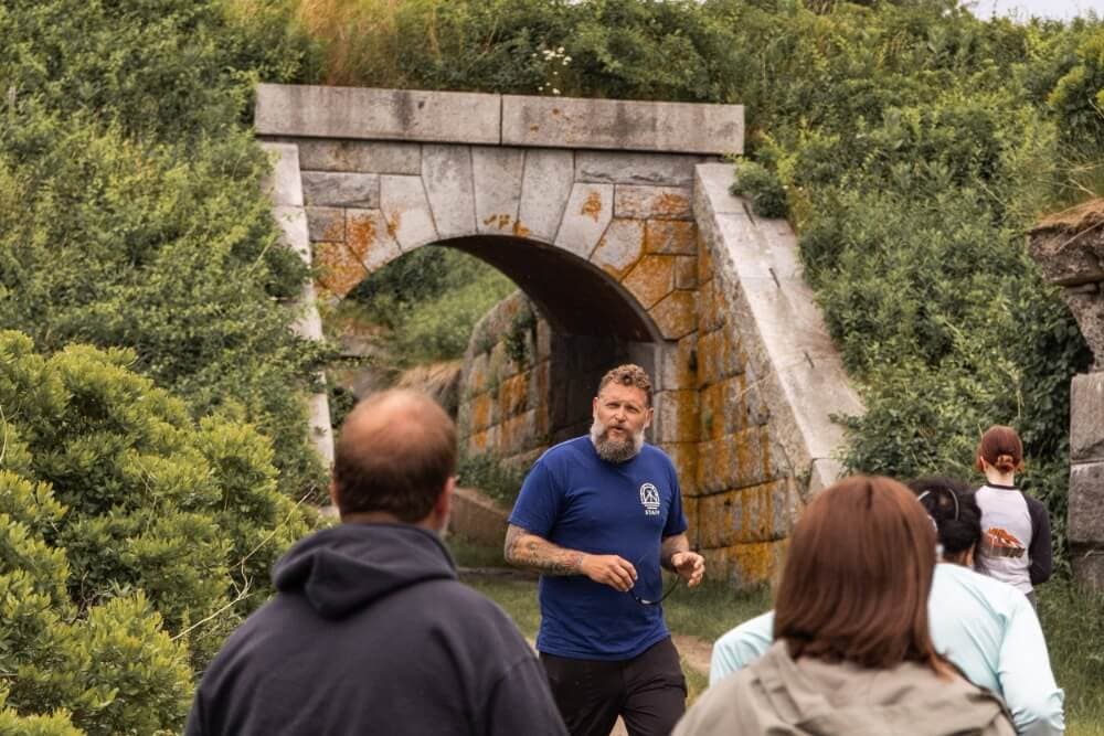 A group of people listens to a guide near a stone archway covered in greenery.