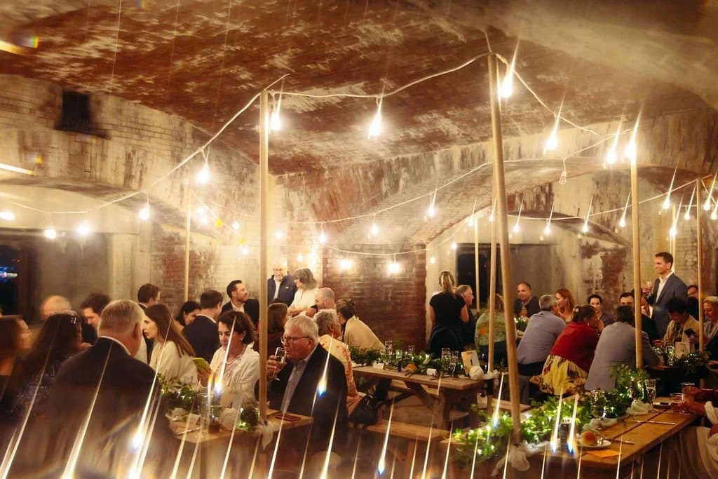 A lively dinner party takes place in a warmly lit, rustic venue with guests seated at long tables.