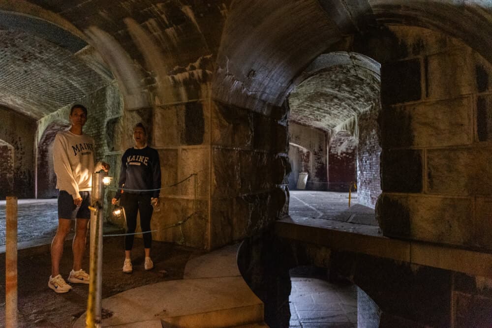 Two people stand in a dimly lit, arched stone chamber holding flashlights.