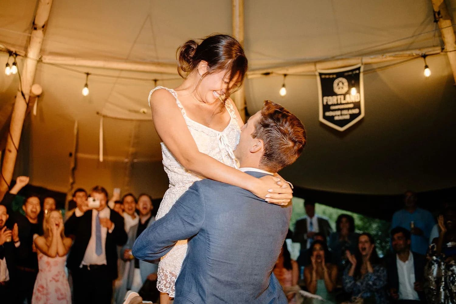 A groom joyfully lifts his bride during their wedding celebration, surrounded by cheering guests.
