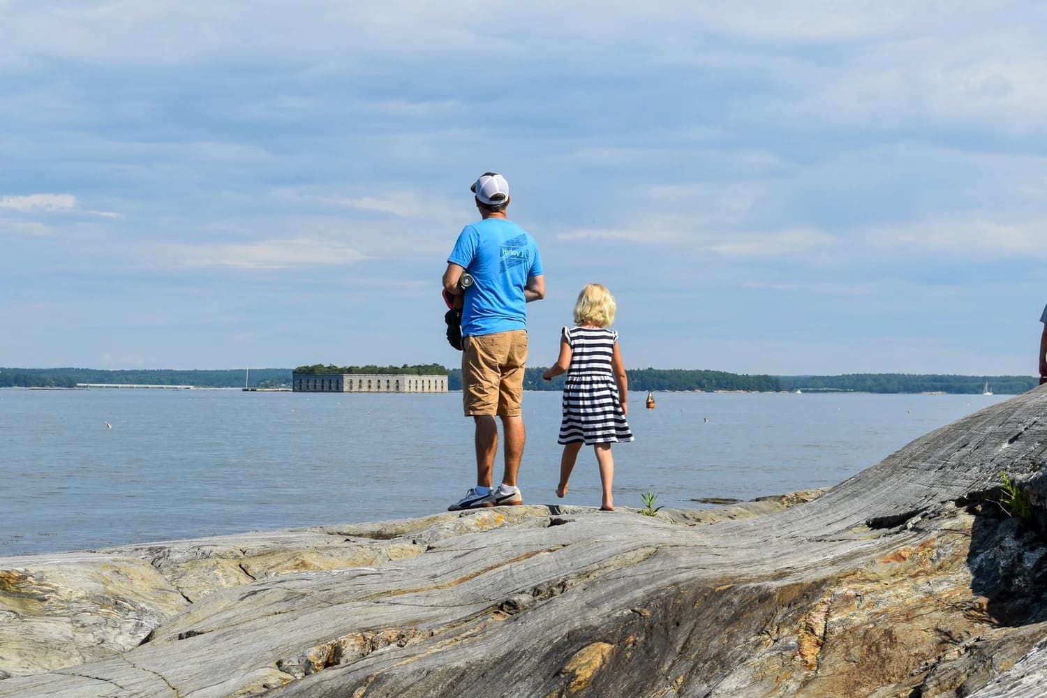 A man and a child stand on a rocky shore, gazing out at a body of water under a cloudy sky.