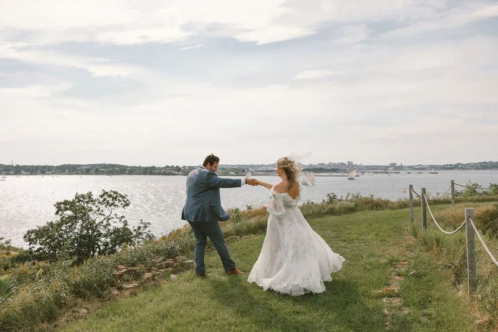 A bride and groom dance playfully on a grassy shore with water in the background.