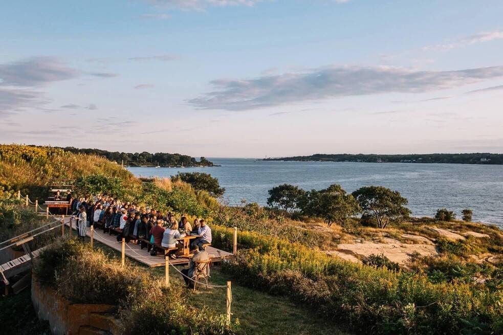 A large group of people gathers at a seaside table during sunset.