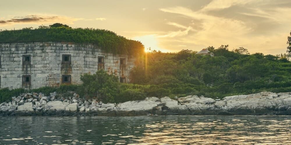 A sunlit, overgrown stone ruin sits on a rocky shore beside calm water.
