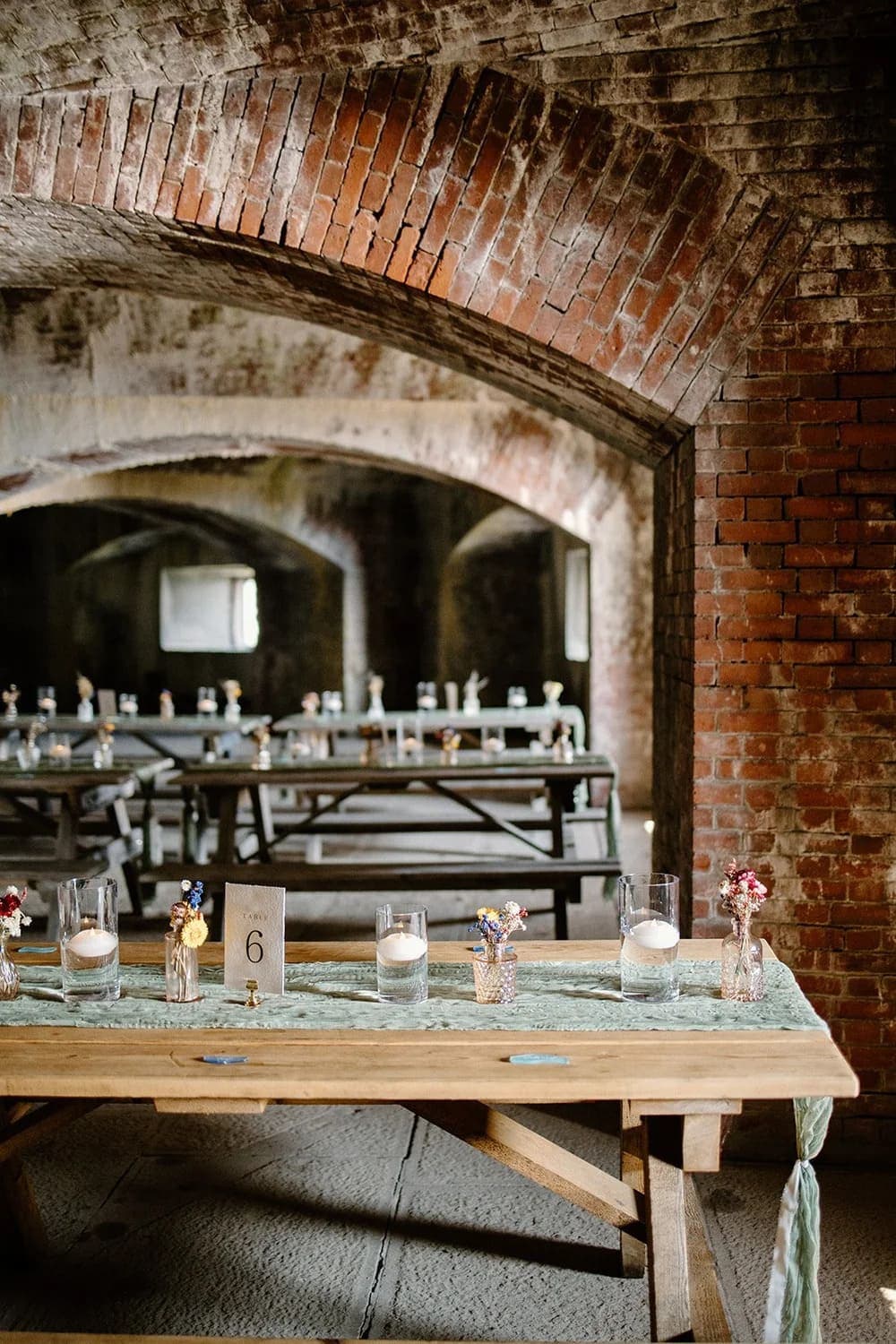 A rustic venue setup with wooden tables, decorated with flowers and candles, under an arched brick ceiling.