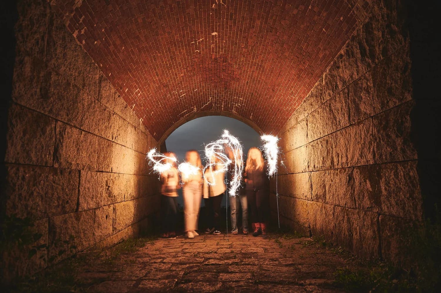 People holding sparklers create light trails inside a dark brick tunnel.