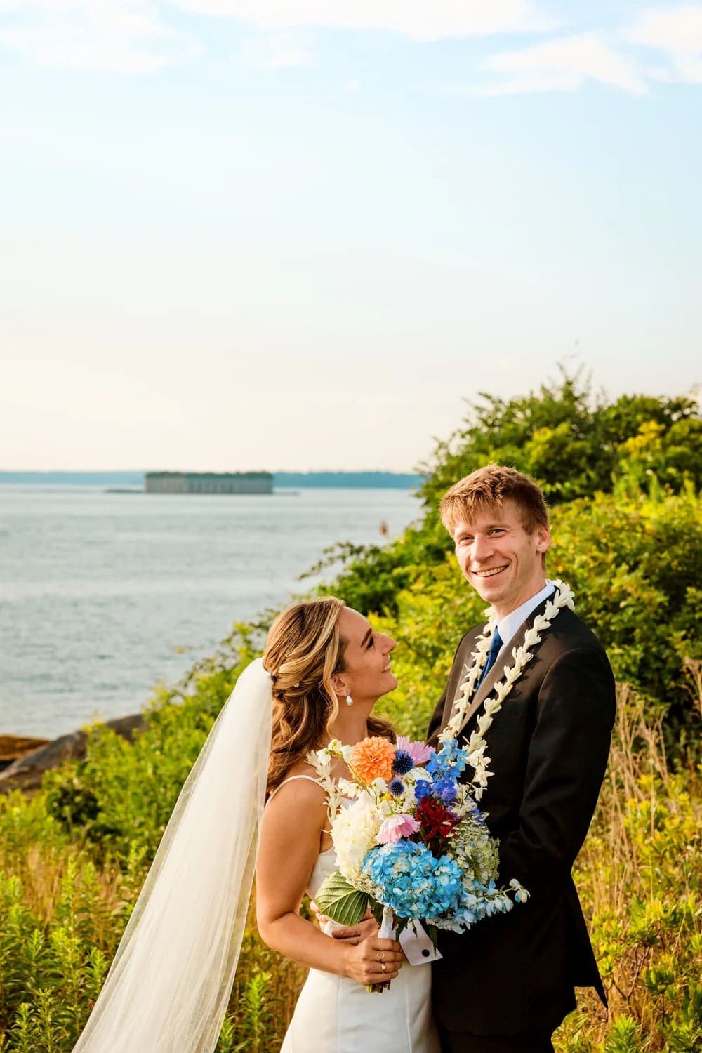A bride and groom smile at each other, with the ocean and greenery in the background.