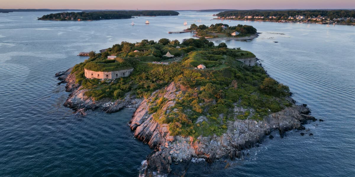 Aerial view of a small, lush island with historical fort ruins surrounded by water at sunset.
