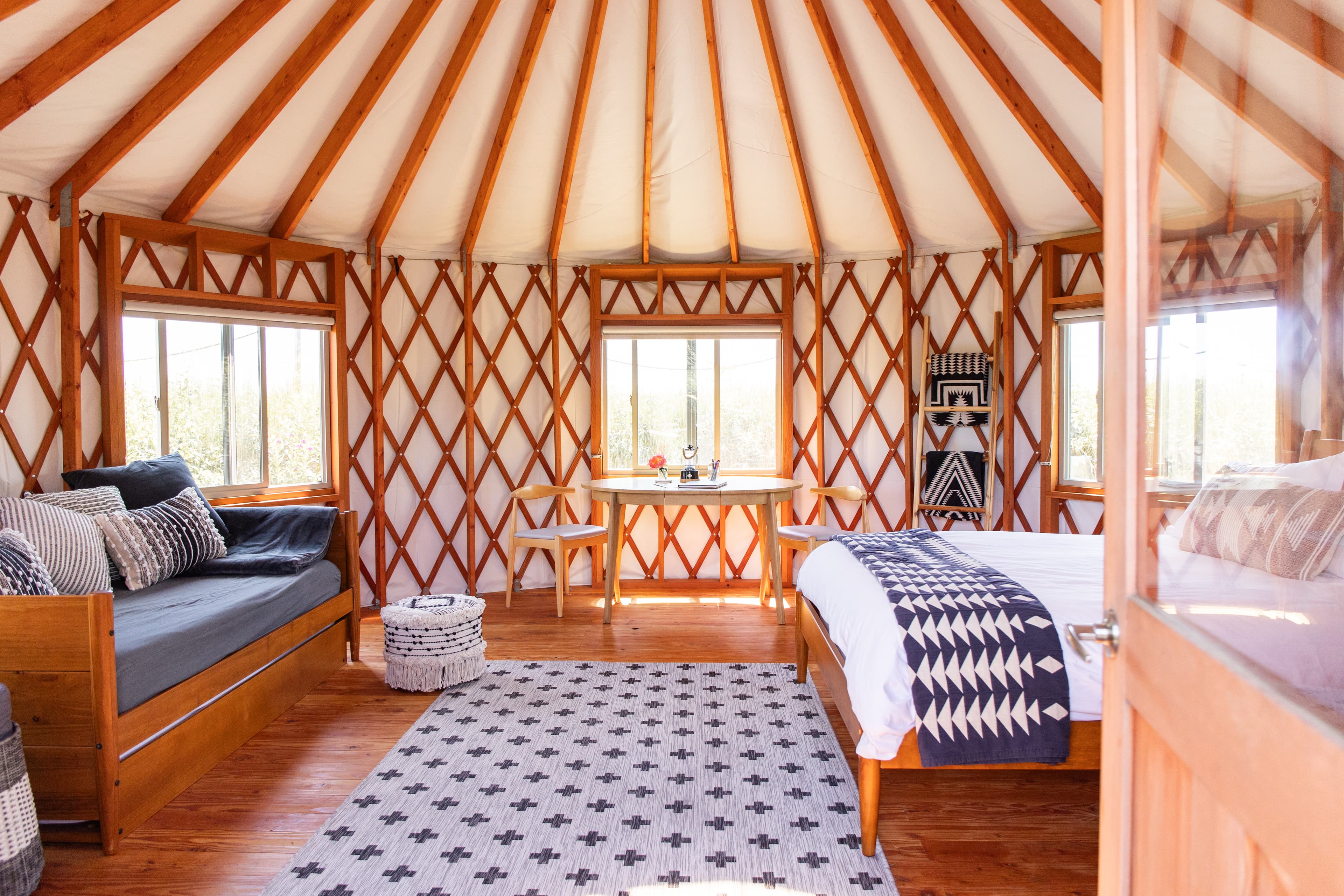 A cozy, circular yurt interior featuring wooden beams, a bed, a daybed, and large windows.