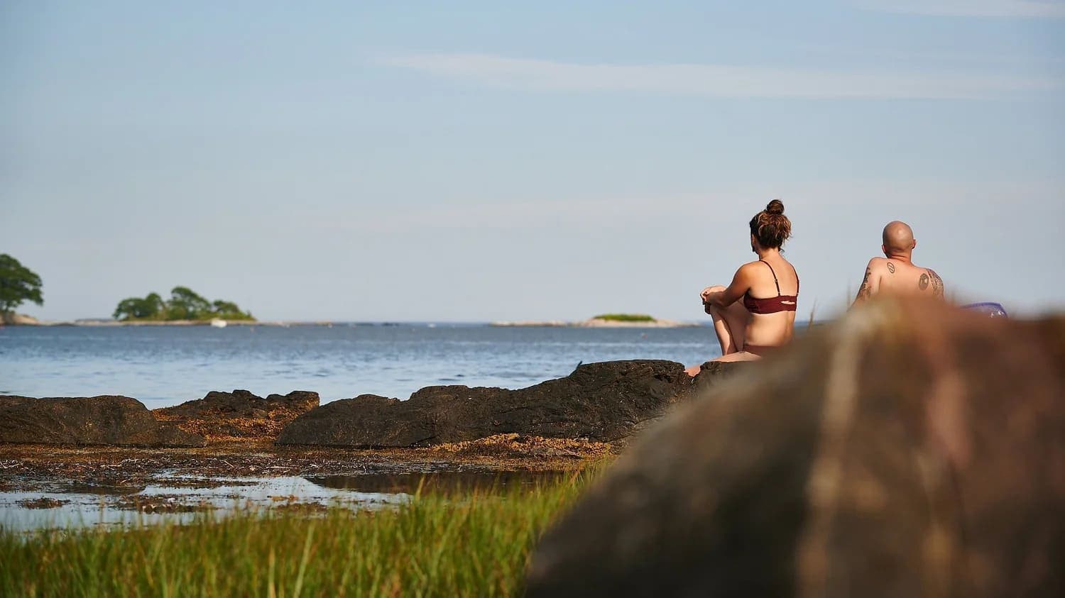 A woman and a man sit on rocks by a serene body of water, gazing at the horizon.