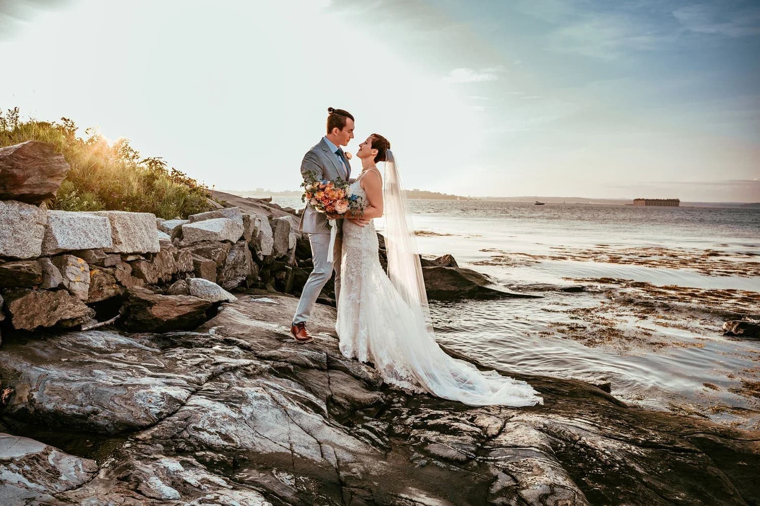 A couple in wedding attire stands on a rocky shoreline, sharing a romantic moment with the ocean in the background.