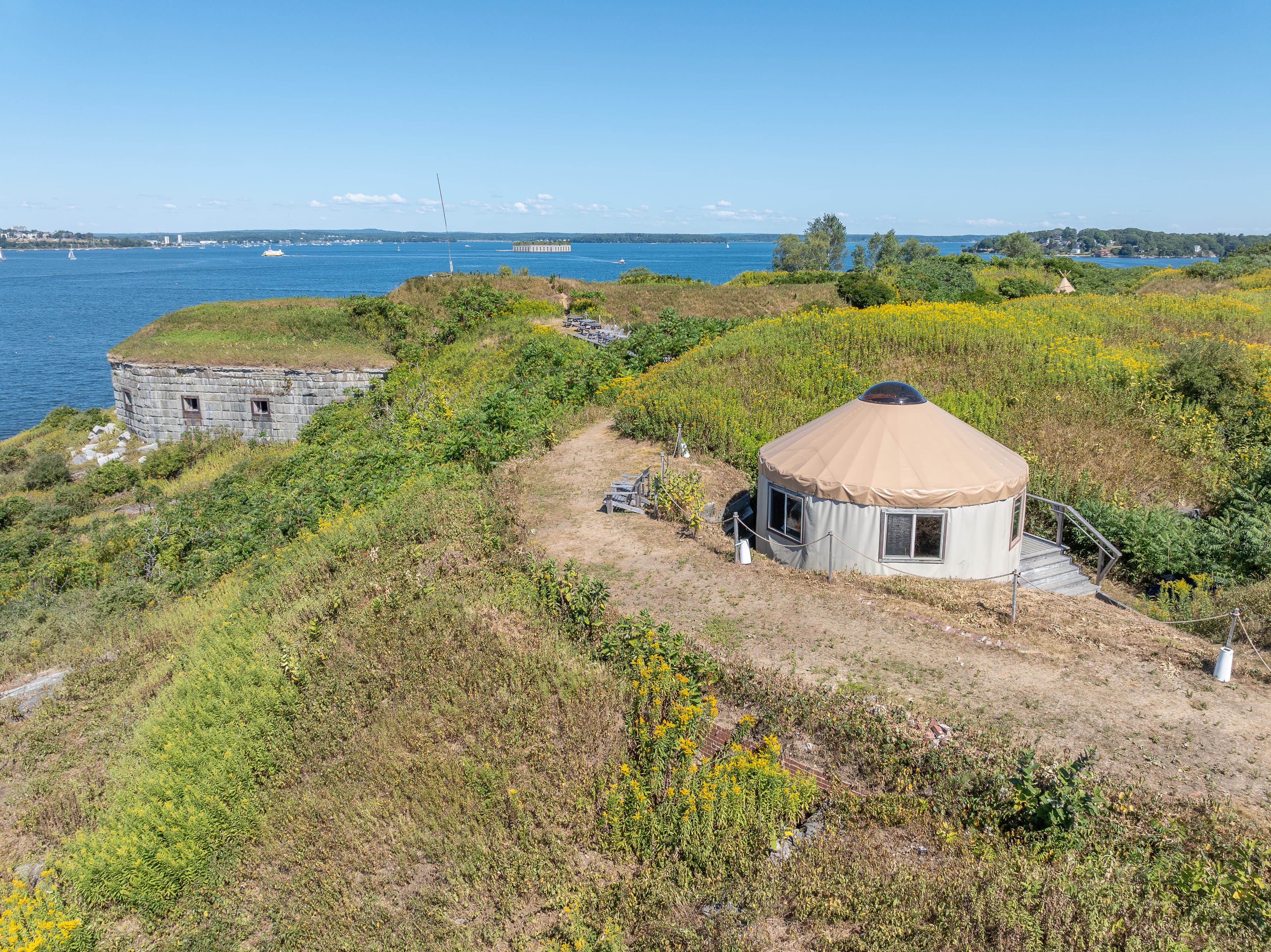 A yurt sits on a hillside overlooking a tranquil body of water and lush greenery.