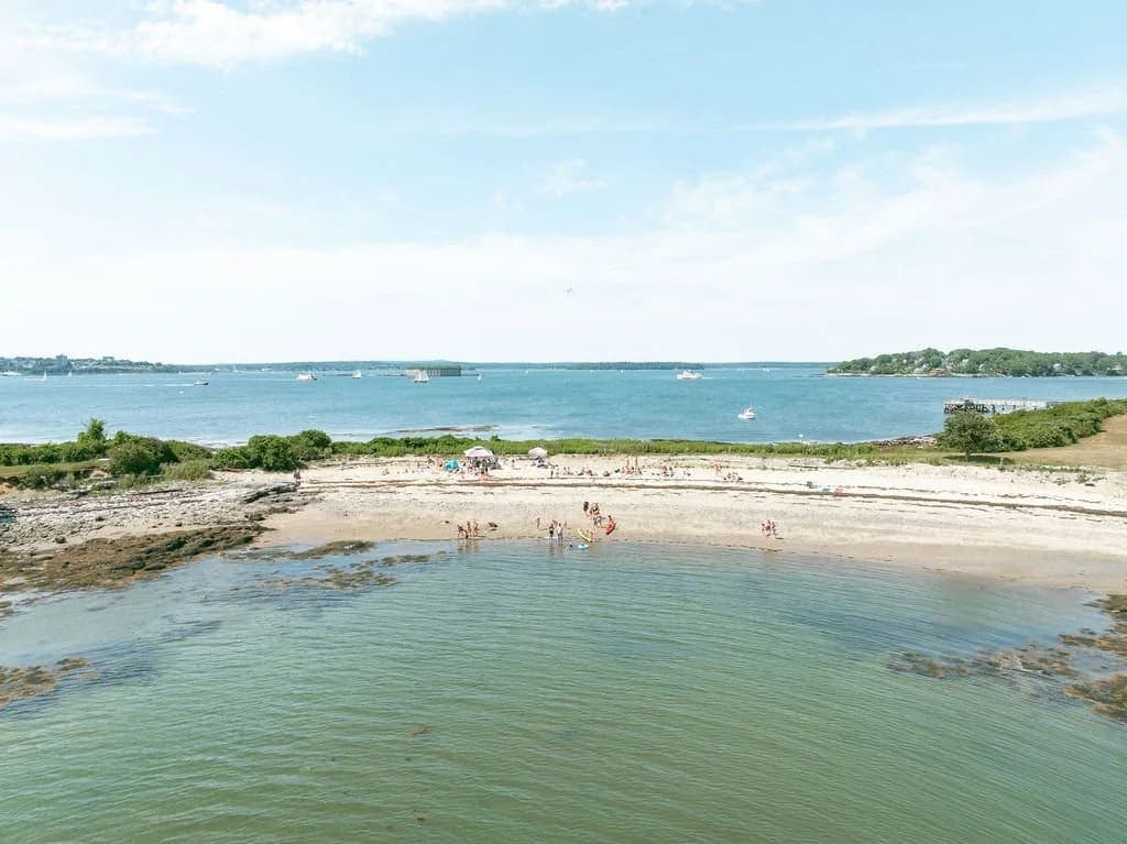A scenic view of a beach with people enjoying the water, surrounded by boats and greenery under a clear sky.