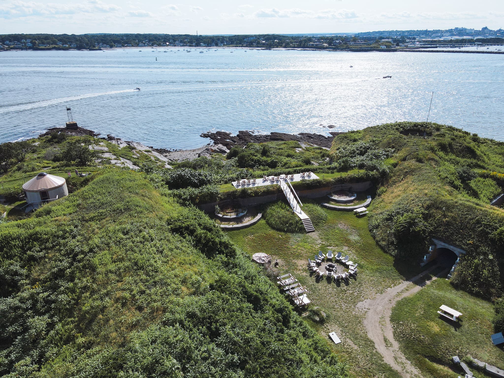 Aerial view of a coastal area featuring grassy mounds, a gathering space, and boats on the water.