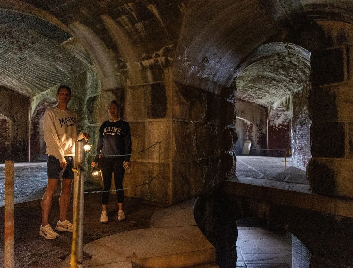 Two individuals stand in a dimly lit, historic stone tunnel, holding flashlights.