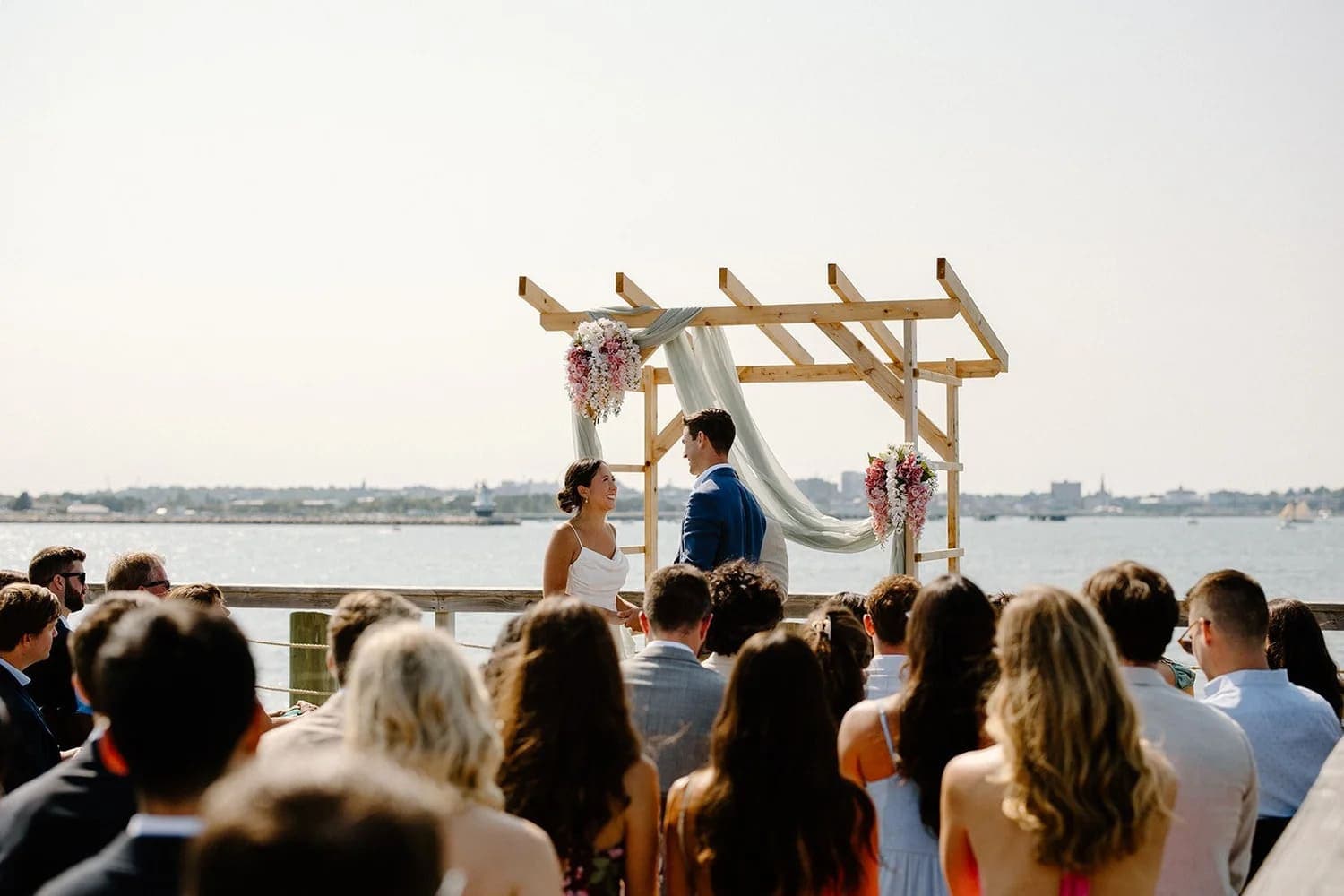 A bride and groom exchange vows under an arch by the water, surrounded by guests.