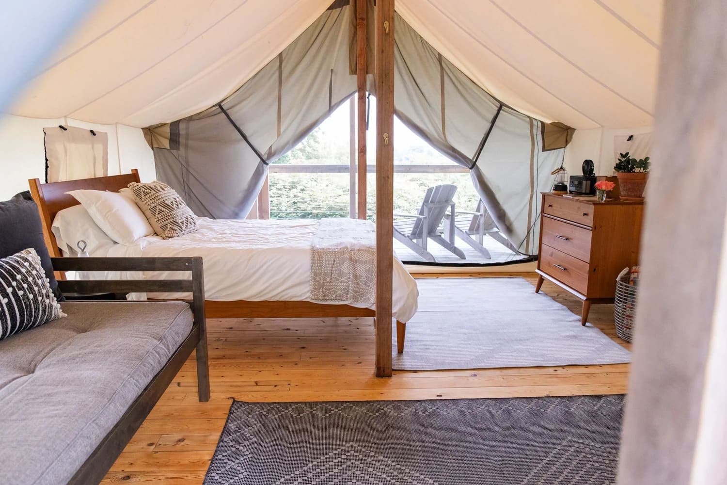 Interior of a cozy glamping tent featuring a bed, a couch, a dresser, and large windows overlooking an outdoor deck.