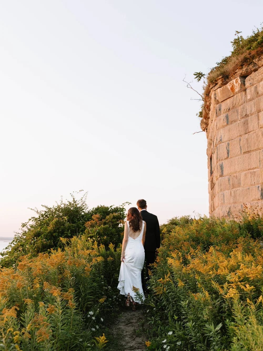 A couple walks hand-in-hand along a path surrounded by wildflowers near a stone structure.