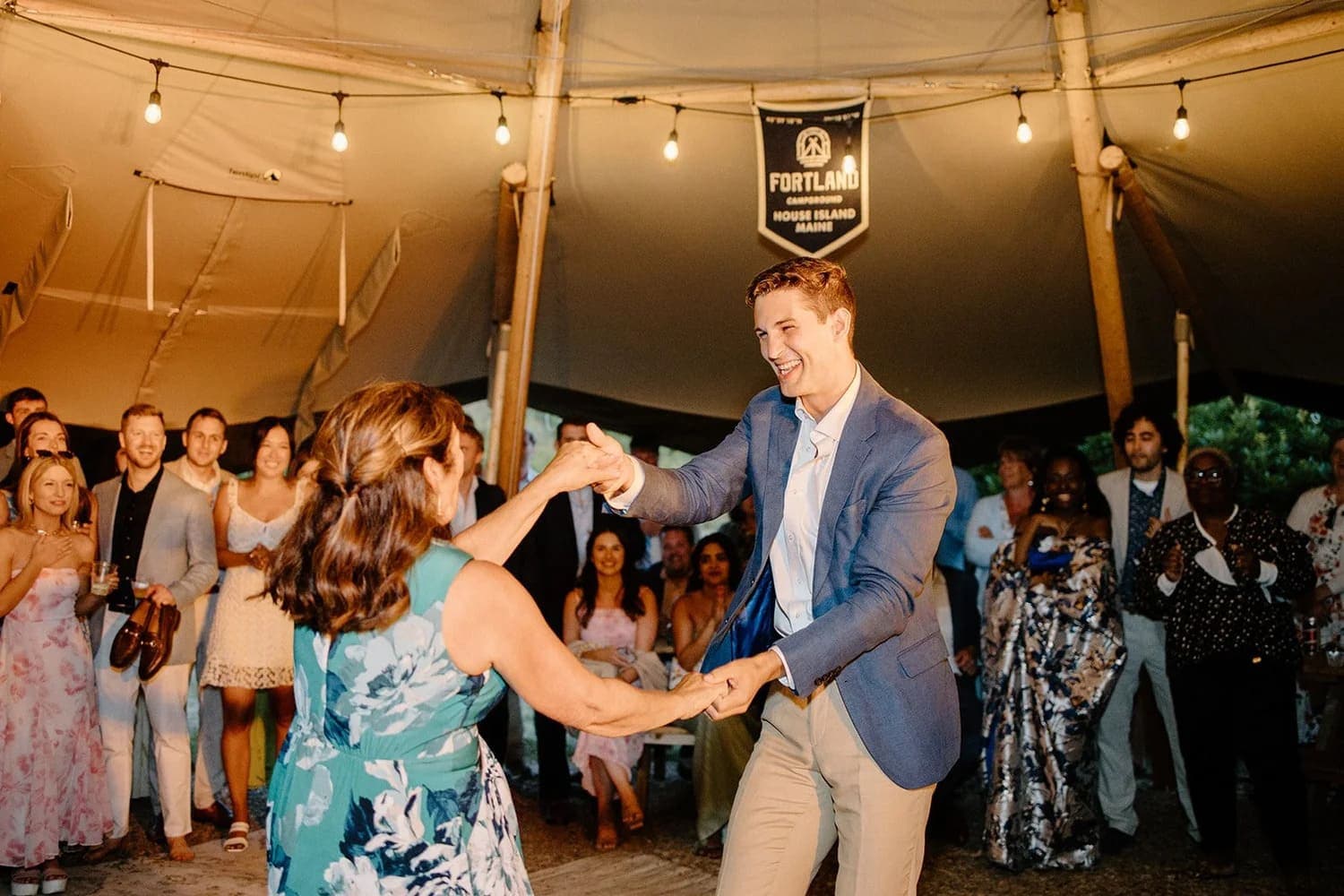 A man in a suit dances with a woman in a floral dress, surrounded by smiling guests in a festive tent setting.