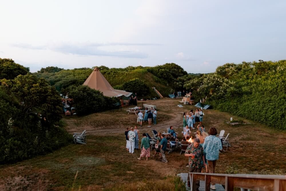 A group of people gathers around a campfire in a lush, green outdoor setting at dusk, with a large tent in the background.