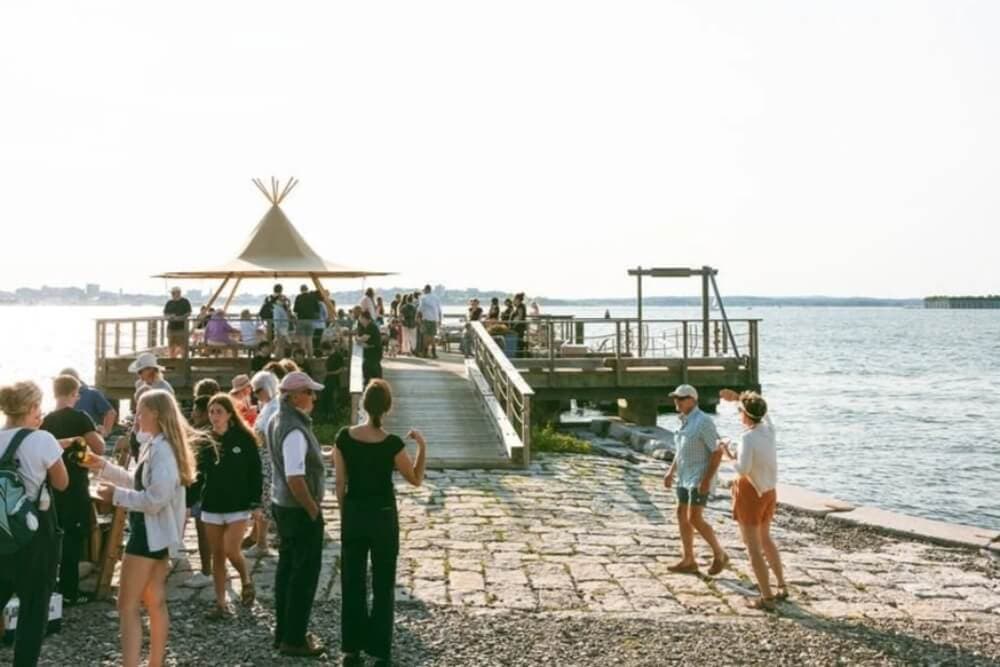 A lively gathering of people on a pier with a view of the water and a teepee structure in the background.