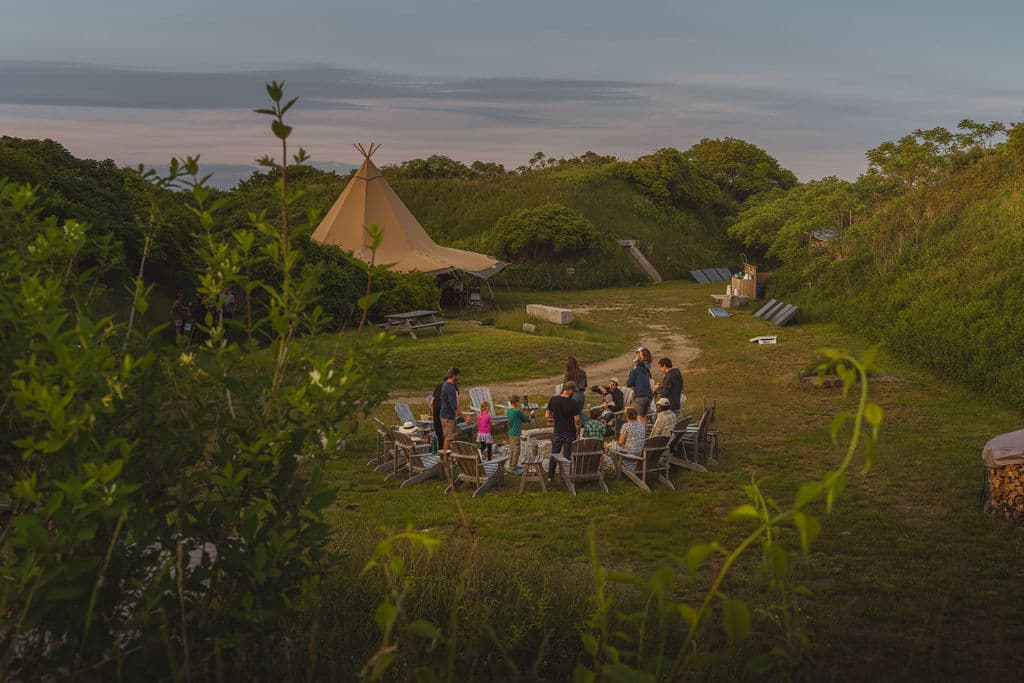 A group of people gathers around a fire pit in a grassy area with a teepee in the background at dusk.