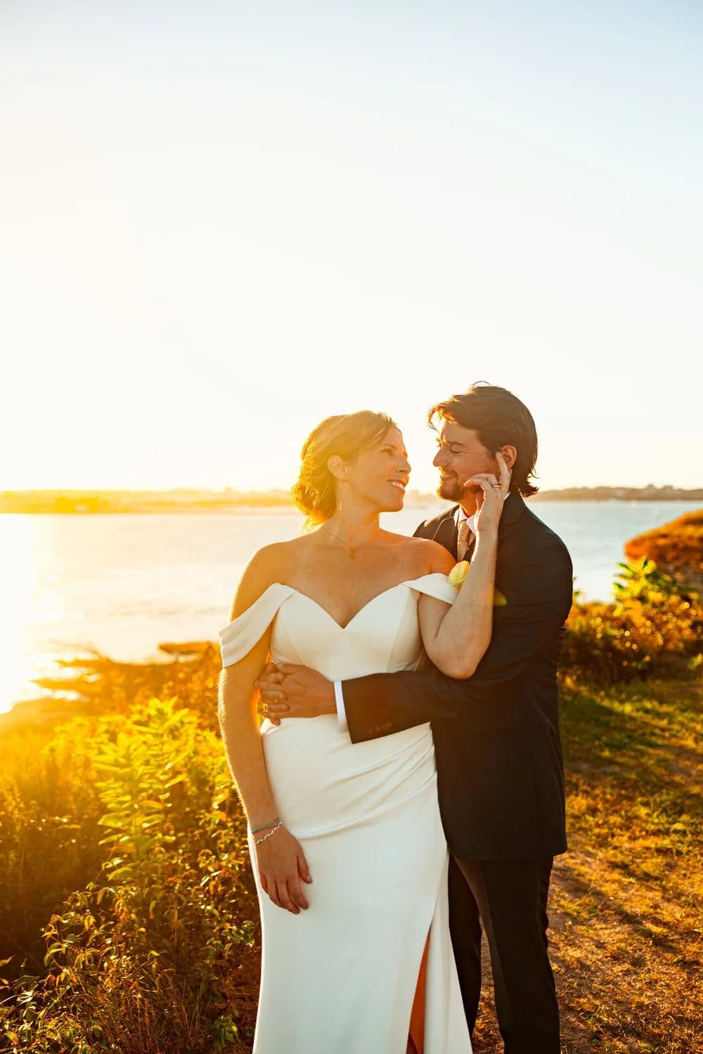 A couple shares a tender moment at sunset, with the bride in a flowing white gown and the groom in a black suit.