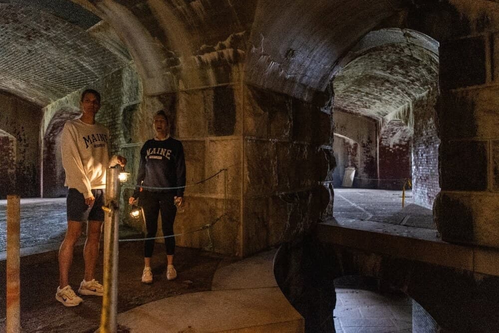 Two people stand in a dimly lit, historical stone chamber holding a lantern.