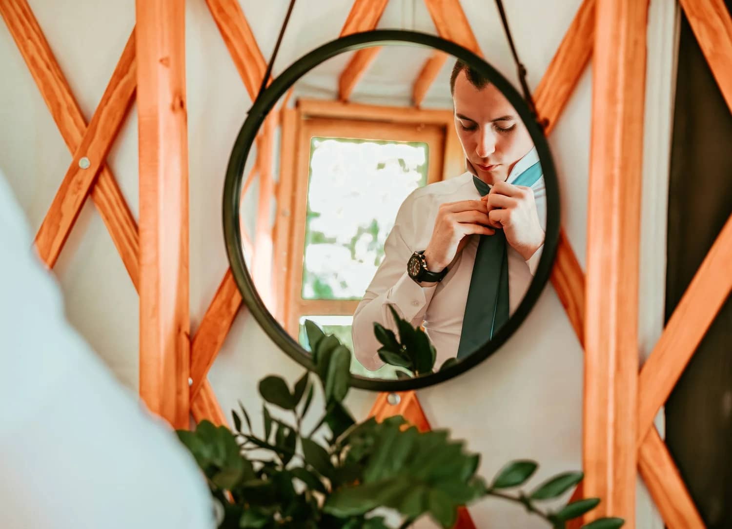 A man adjusts his tie while looking in a round mirror surrounded by wooden accents.