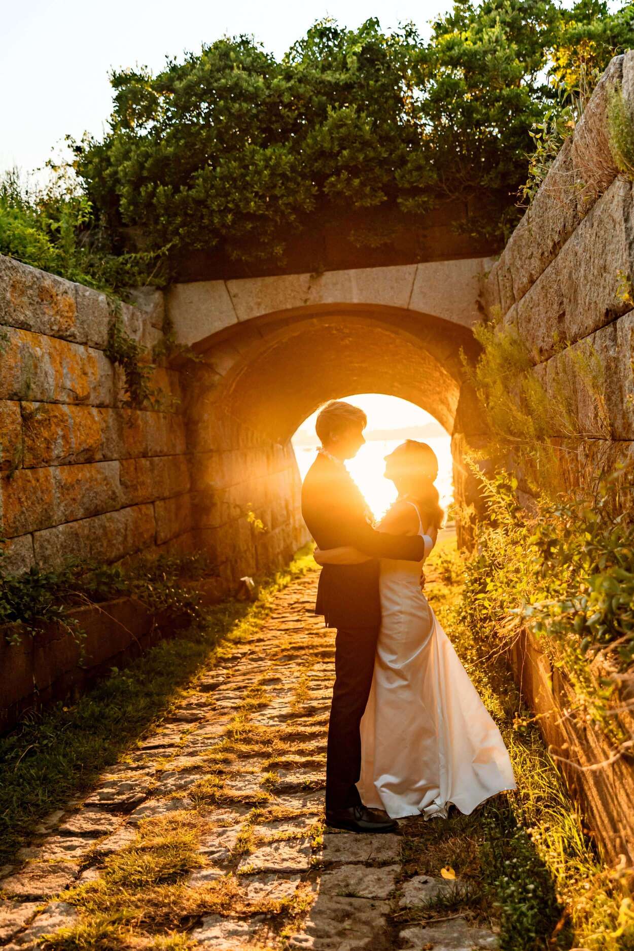 A couple embraces in silhouette under a stone archway at sunset.