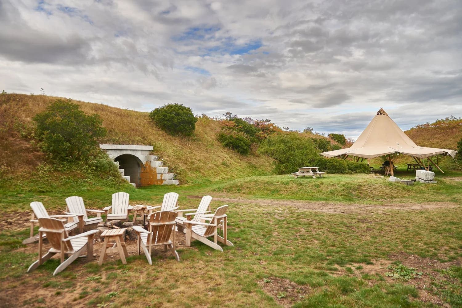 A campsite featuring a tent, picnic table, and Adirondack chairs surrounded by grassy hills under a cloudy sky.